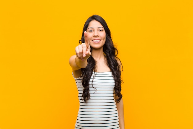 Young pretty woman smiling proudly and confidently making number one pose triumphantly, feeling like a leader against orange wall