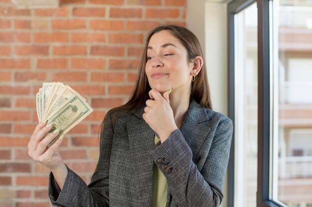 Foto giovane bella donna sorridente con un'espressione felice e sicura con la mano sul mento banconote in dollari conc