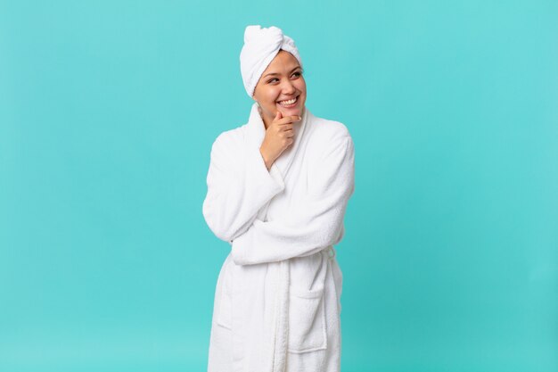 Young pretty woman smiling with a happy, confident expression with hand on chin and wearing bath robe after shower