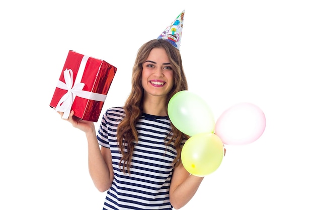 Young pretty woman in stripped T-shirt and celebration cap holding red present and colored balloons on white background in studio