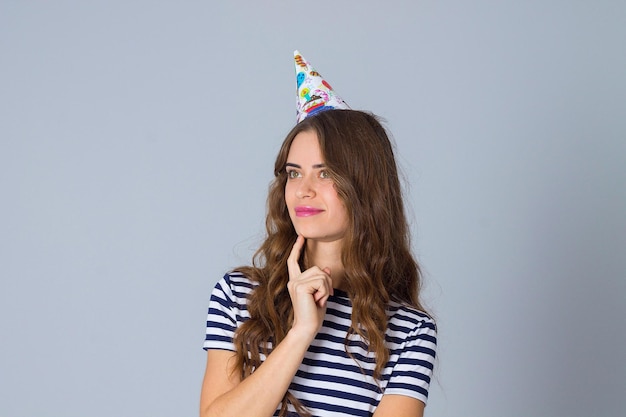 Young pretty woman in stripped T-shirt and celebration cap thinking on grey background in studio