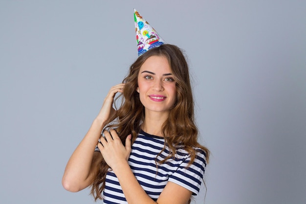 Young pretty woman in stripped T-shirt and celebration cap touching her long hair on grey background in studio