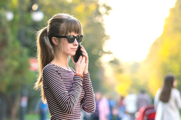 暖かい秋の日に屋外の街の通りで携帯電話で話している若いきれいな女性。