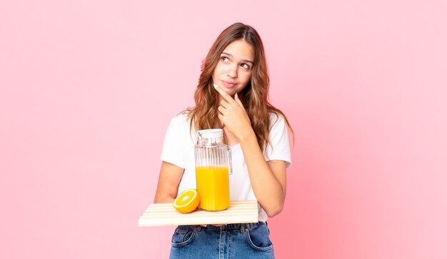 Young pretty woman thinking, feeling doubtful and confused and holding a tray with an orange juice