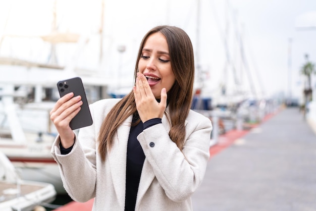 Giovane donna graziosa che utilizza il telefono cellulare all'aperto con espressione facciale sorpresa e scioccata