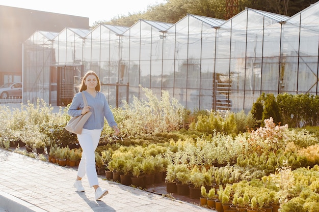 Молодая красивая женщина идет по улице возле больших теплиц Женщина идет покупать зеленые растения для украшения Концепция ландшафтного дизайна