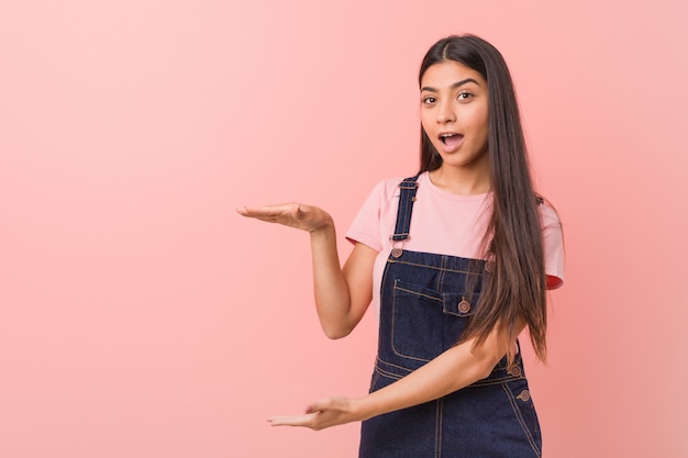 데님 dungaree를 입고 젊은 예쁜 여자는 충격과 손 사이에 뭔가 들고 놀라 울