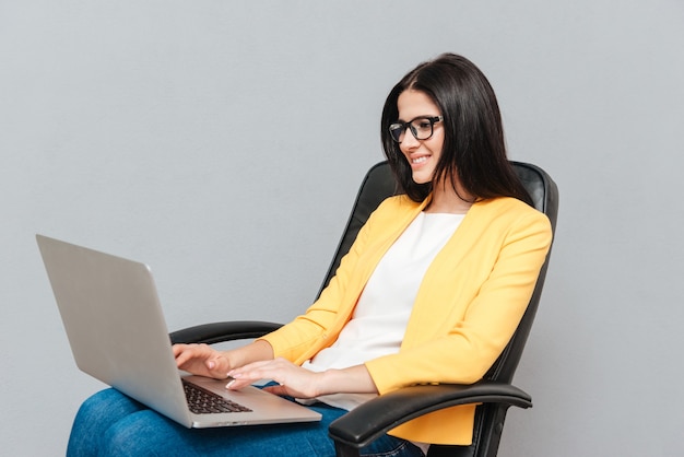 Young pretty woman wearing eyeglasses and dressed in yellow jacket sitting on office chair while using laptop over grey surface.