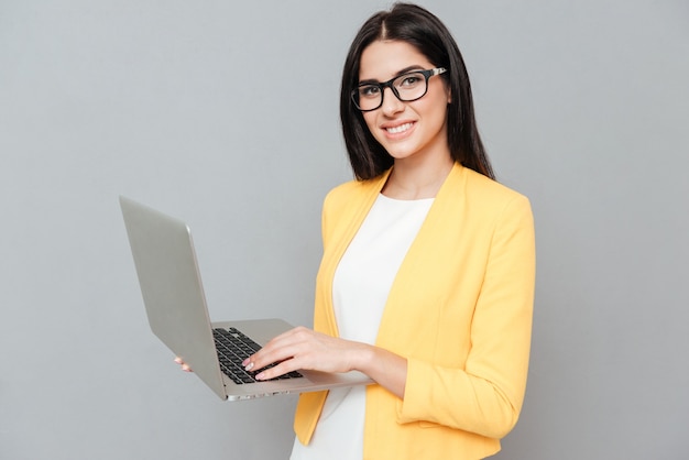 Young pretty woman wearing eyeglasses and dressed in yellow jacket using laptop over grey surface.