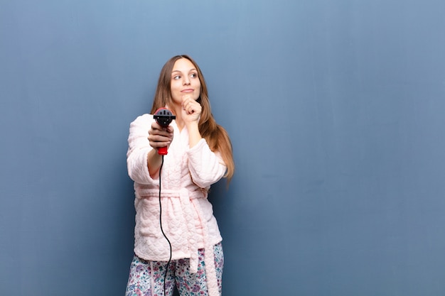 Young pretty woman wearing pajamas and holding a hairdresser against blue wall with a copyspace