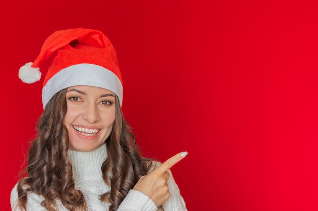サンタクロースの帽子をかぶった美しい若い女性が微笑み ⁇ 空の赤い背景に指を指しています ⁇ 