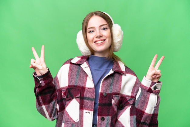 Young pretty woman wearing winter muffs over isolated chroma key background showing victory sign with both hands