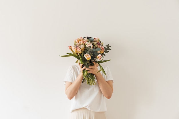 Young pretty woman in white blouse holding tulip flowers bouquet in hands against white wall