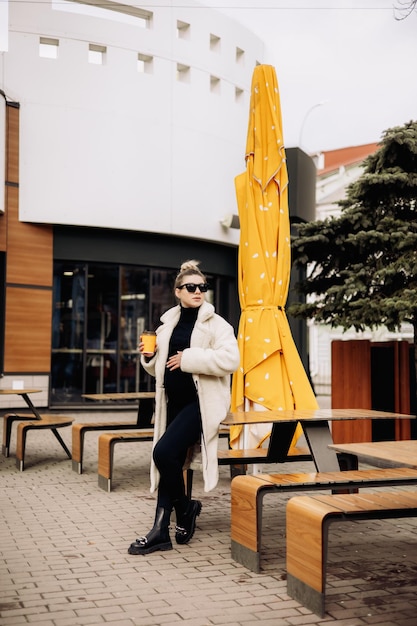 A young pretty woman in a white fur coat and sunglasses with coffee walks around the city Stylish young girl