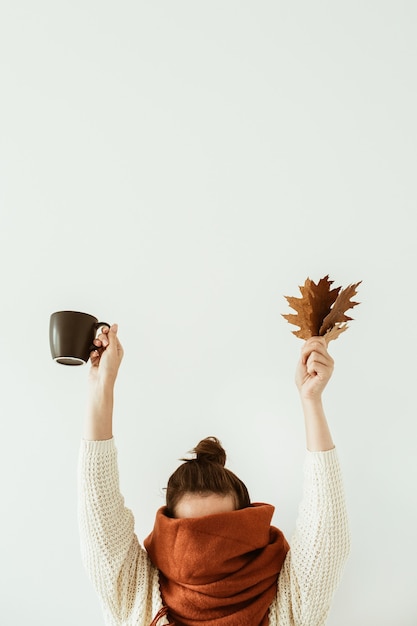 Young pretty woman in white sweater with brown scarf hold mug and fall leaves in hands on white