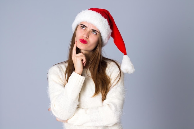 Young pretty woman in white sweater with red christmas hat wondering on grey background in studio