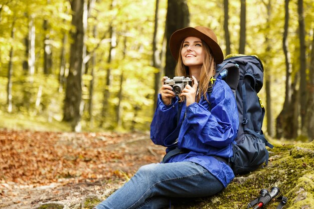 Photo young pretty woman with backpack and camera wearing hat hiking in autumn forest in mountains she making photos of forest trekking recreation and healthy lifestyle concept