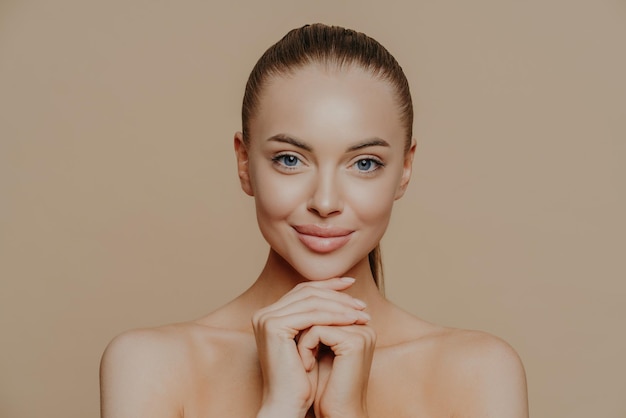 Photo young pretty woman with beauty skin and combed hair, keeps hands under chin, has healthy complexion after face purification, soft well cared body, isolated over studio wall. facial treatment