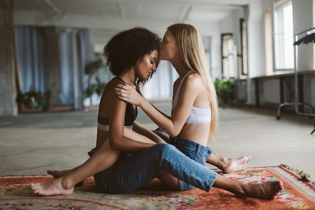 Photo young pretty woman with blond hair dreamily kissing in forehead nice african american woman with dark curly hair while sitting on carpet together at home