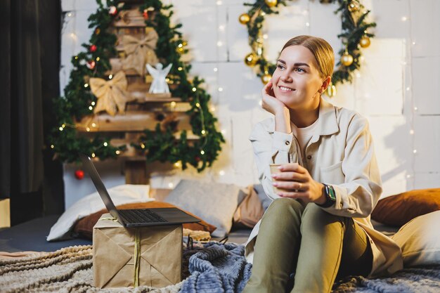 Young pretty woman with blond long hair in cozy clothes with\
coffee in hands using laptop watching movie and smiling on bed in\
room with christmas tree at home new year holidays