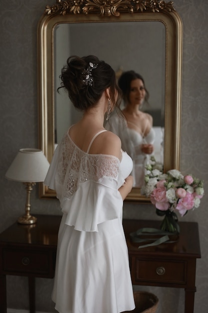 A young pretty woman with bridal coiffure in lace peignoir is posing in front of the mirror a beauti...