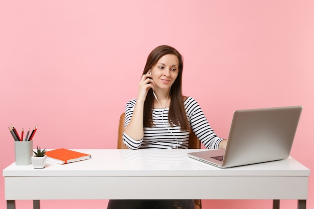 ビデオ通話を作る音楽を聞くイヤホンを持つ若いきれいな女性は、現代のPCラップトップでオフィスに座って仕事をする