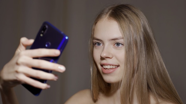 Giovane bella donna con facce buffe fa selfie mentre prende selfie su smartfone su sfondo grigio