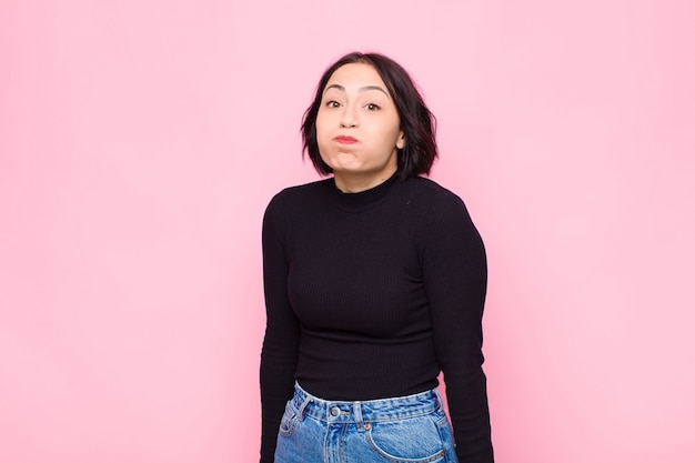 Young pretty woman with a goofy, crazy, surprised expression, puffing cheeks, feeling stuffed, fat and full of food against pink wall