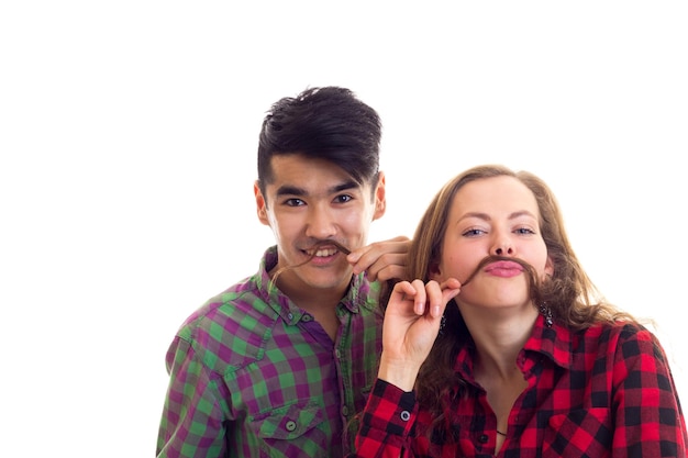 Young pretty woman with long chestnut hair and young handsome man with dark hair in plaid shirts making moustache of hair on white background in studio