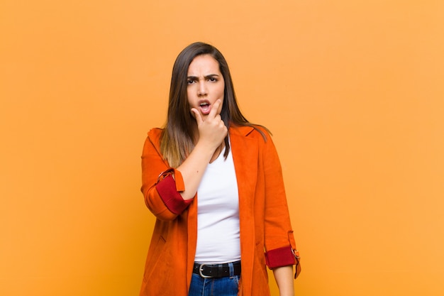 Young pretty woman with mouth and eyes wide open and hand on chin, feeling unpleasantly shocked, saying what or wow isolated against orange wall