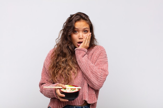 Young pretty woman with noodles feeling shocked and scared, looking terrified with open mouth and hands on cheeks