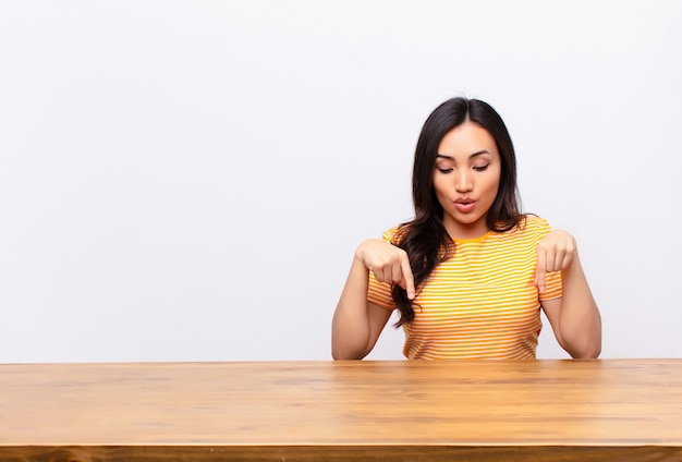 Young  pretty woman with open mouth pointing downwards with both hands, looking shocked, amazed and surprised against flat wall