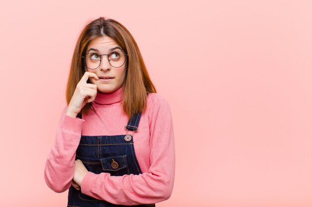 Young pretty woman with surprised, nervous, worried or frightened look, looking to the side towards copy space over pink wall