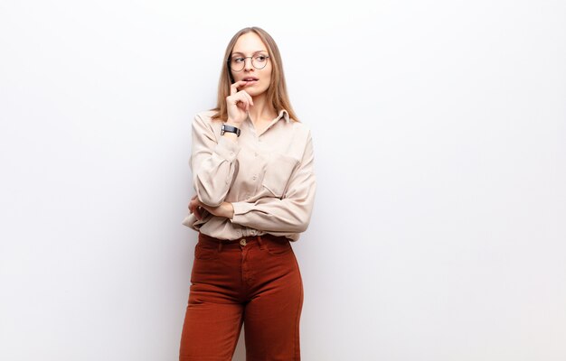 Young pretty woman with surprised, nervous, worried or frightened look, looking to the side towards copy space over white wall