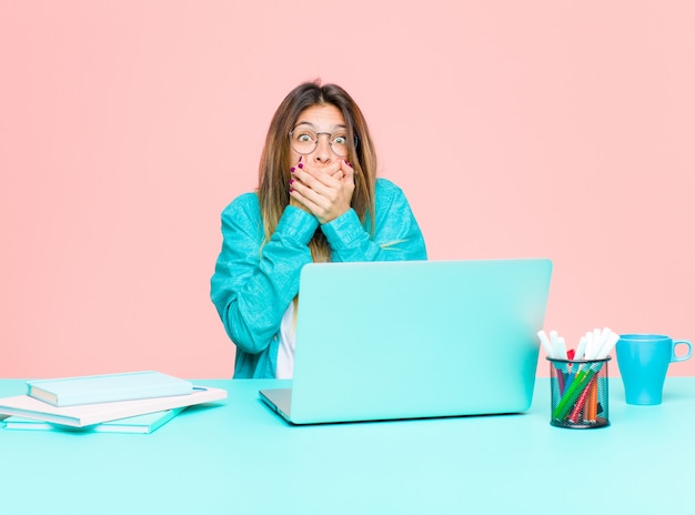 Young pretty woman working with a laptop covering mouth with hands with a shocked, surprised expression, keeping a secretsaying oops