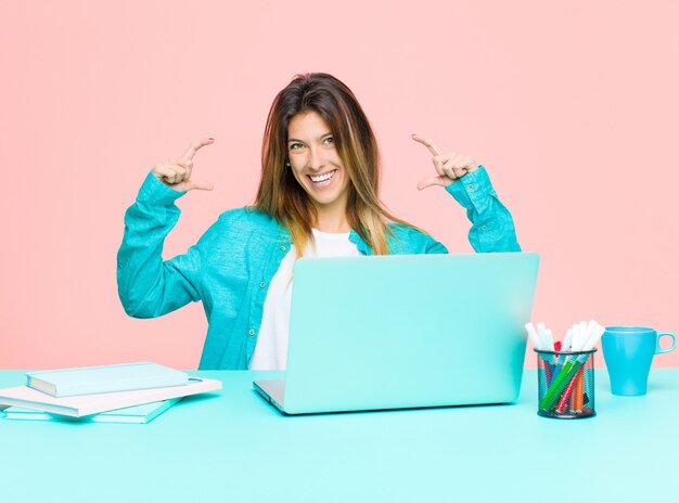 Young pretty woman working with a laptop framing or outlining own smile with both hands, looking positive and happy, wellness concept