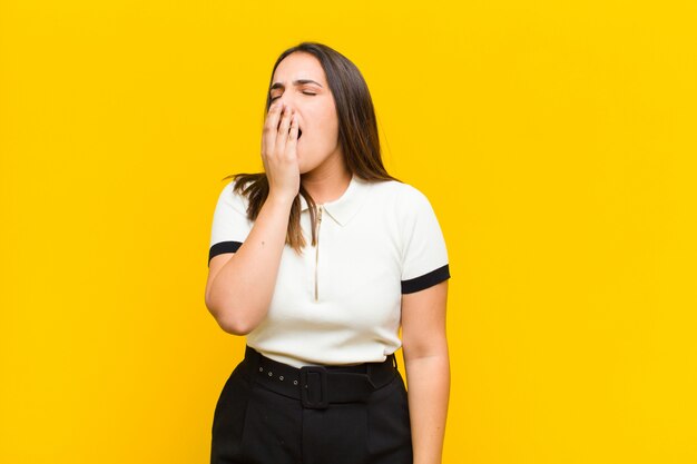 Young pretty woman yawning lazily early in the morning, waking and looking sleepy, tired and bored isolated against orange wall