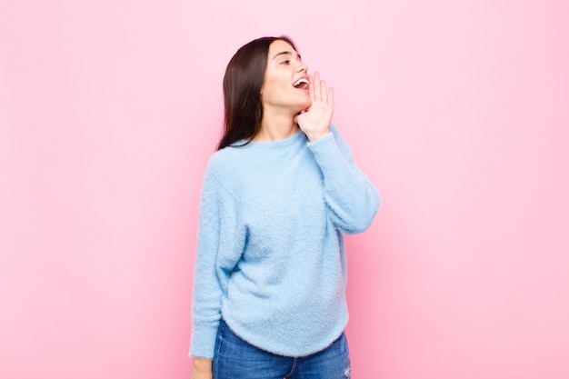 Young pretty woman yelling loudly and angrily to copy space on the side, with hand next to mouth against pink wall