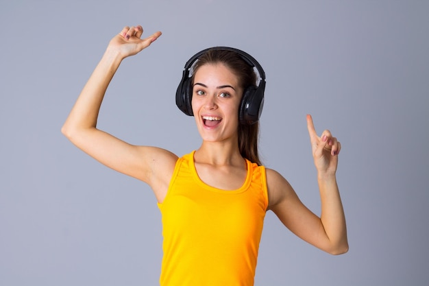 灰色の背景に黒いヘッドフォンで音楽を聴いている黄色のTシャツの若いきれいな女性