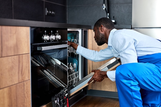 young professional african american handyman in blue overalls uniform is going to repair dishwasher, confident contractor examining it before repair, looking serious, side view portrait.