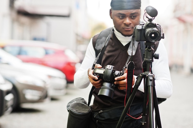 Giovane videografo afroamericano professionista che tiene una fotocamera professionale con attrezzatura pro treppiede cameraman afro che indossa duraq nero che realizza un video