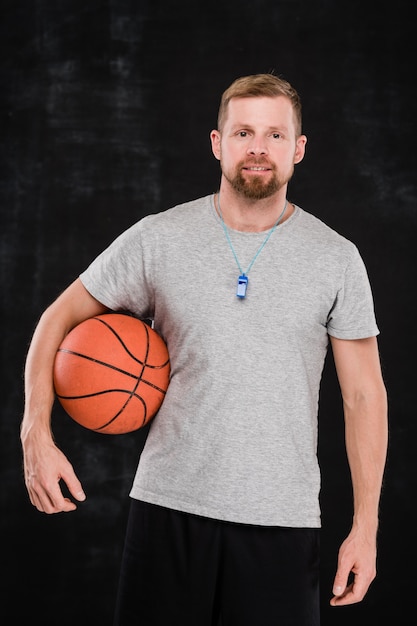 Young professional basketball trainer with ball standing in front of camera against black background