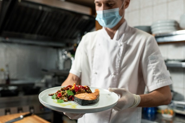 Giovane chef professionista in uniforme bianca e maschera protettiva e guanti che tengono piatto con salmone fritto e contorno di verdure in cucina