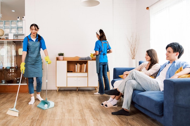 Young professional cleaning service women worker working in the house One sweeps broomsticks on the wooden floor and the other cleans the low cabinet while a couple of owners sit on the sofa