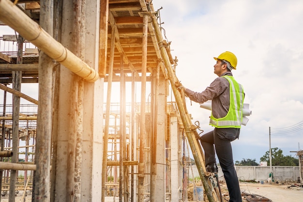 Young professional engineer worker in protective helmet and blueprints paper on hand working on ladder at the house building construction site