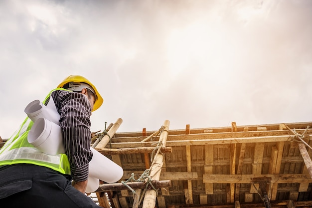 Young professional engineer worker in protective helmet and blueprints paper on hand working on ladder at the house building construction site