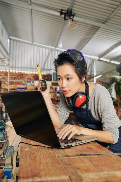 Young professional female carpenter with laptop checking dimensions of furniture item she is going to make in workplace