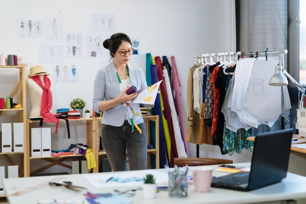 스케치와 바느질 액세서리를 들고 사무실에서 걷는 젊은 전문 여성 양장점. 새로운 가을 시즌 옷을 위한 보라색 키트를 선택하는 스튜디오의 여성 패션 디자이너