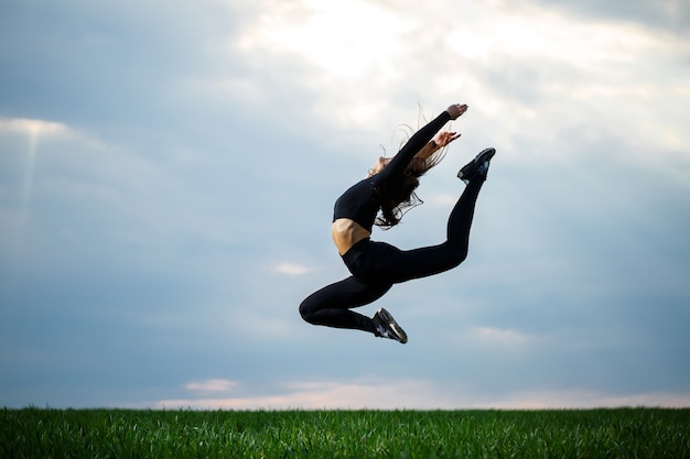 젊은 전문 체조 선수가 푸른 하늘을 배경으로 자연 속에서 뛰어오르고 있습니다. 검은색 상의와 검은색 레깅스를 입은 여자 운동선수는 곡예 운동을 한다