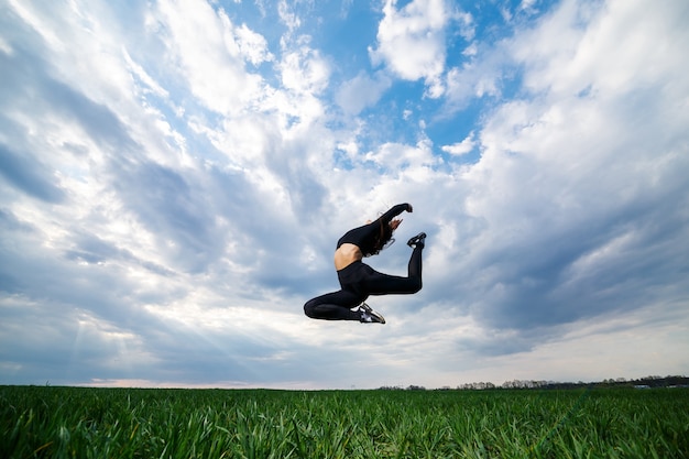 Young professional gymnast is jumping in nature against the blue sky. Girl athlete in a black top and black leggings does acrobatic exercises